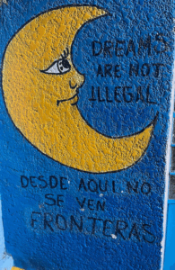 A mural of a yellow crescent moon on a blue background with text in English and Spanish that reads "dreams are not illegal / desde aqui no se ven fronteras"