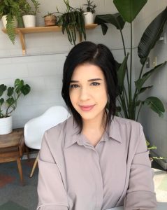 Smiling woman with dark hair sitting with plants on shelf and white walls behind