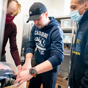 Three people looking at items inside a cardboard box; two wearing masks and one with a black cap and navy hoodie.