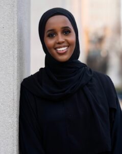 Muna smiling while leaning against gray wall.