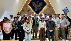 A group of 21 people wearing flat caps, posing indoors beneath a large wall decoration of a phoenix.