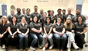 A group of 19 people in matching dark gray shirts, posing indoors in front of signs labeled "BANK STOCK," "A to K," and "L to Z."