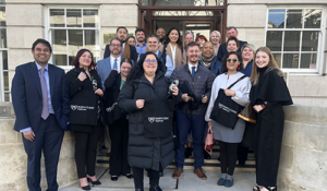 A diverse group of people posing outside a building with arched windows, holding black tote bags.
