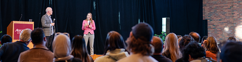 Two speakers on a stage with an audience seated in front.