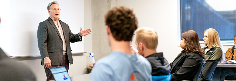A man speaks to students in a classroom, gesturing with his hand.