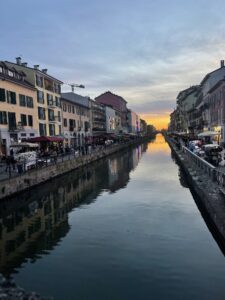 A canal runs through a lively city at sunset, with rows of colorful buildings lining both sides. People walk along the sidewalks and gather at outdoor cafés and market stalls, while the sky glows orange near the horizon and reflects in the calm water below.