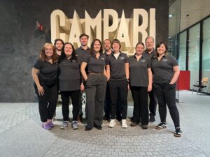 A team of ten people wearing matching grey polo shirts smile for a photo in front of a large, glowing Campari sign.