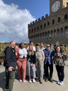 A group of nine adults stands together outdoors in front of a historic stone building with arched windows, decorative shields, and a large clock tower. The group is smiling and dressed in casual to business-casual clothing, many wearing green lanyards. The sky is bright with large clouds, and sunlight casts clear shadows across the terrace where they are gathered.
