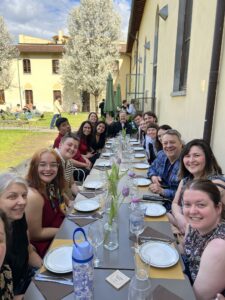 A large group of friends and colleagues smiling around a long outdoor dinner table in a sunny courtyard with spring blossoms in the background.