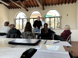 A small group of adults sits around tables in a bright room with high wooden ceilings and large arched windows while an older man stands at the front, gesturing as he speaks. Notebooks, papers, and a pen are visible on the table in the foreground, suggesting a workshop or class setting. Sunlight streams through the windows, illuminating the space and the buildings visible outside.