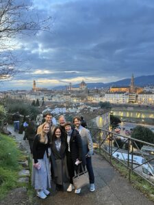 A group of six people stands together smiling on a hillside overlook at dusk, with a panoramic view of a historic city below. The skyline features a large domed cathedral, towers, and softly lit buildings, while dramatic clouds fill the evening sky. Other visitors are scattered along the path, and a railing lines the edge of the overlook.