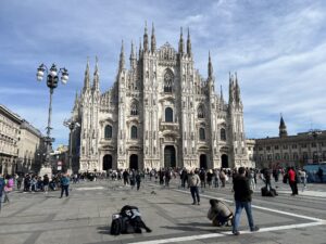 The iconic Gothic Milan Cathedral in Italy, showing its complex facade and numerous spires, with tourists walking through the expansive city square in the foreground.