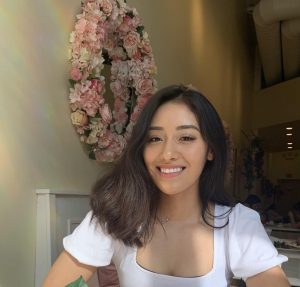 Woman in a white top seated in a restaurant with a floral wreath on the wall next to her