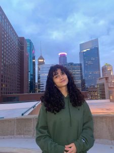 Woman in a green hooded sweatshirt on a rooftop with office buildings and skyscrapers in the background