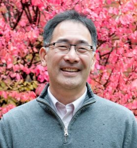 Headshot photo of Andrew Aoki, who is an Asian man wearing a gray pullover over a collared shirt. In the background is a tree with bright pink flowers.