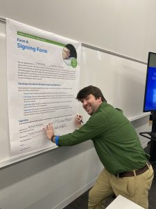 Professor Michael Wentzel, wearing a green shirt and tan pants, signs a giant Green Chemistry Commitment signing form that is taped to a classroom whiteboard.