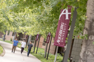 Three students walk under leafy trees on the Augsburg campus towards a maroon banner that reads "Augsburg University"