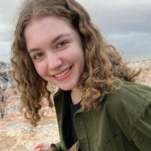 Anna Hudak smiles at the camera. Her hair is curly, and she is outside.