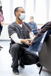 A male nursing student wearing scrubs and a mask kneels down to provide a blood pressure check for a guest at the Augsburg Central Health Commons.