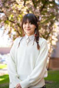 Summer May stands in front of a flowering tree on campus. She is wearing a white collared shirt under a white sweater. Her dark hair is in braids.