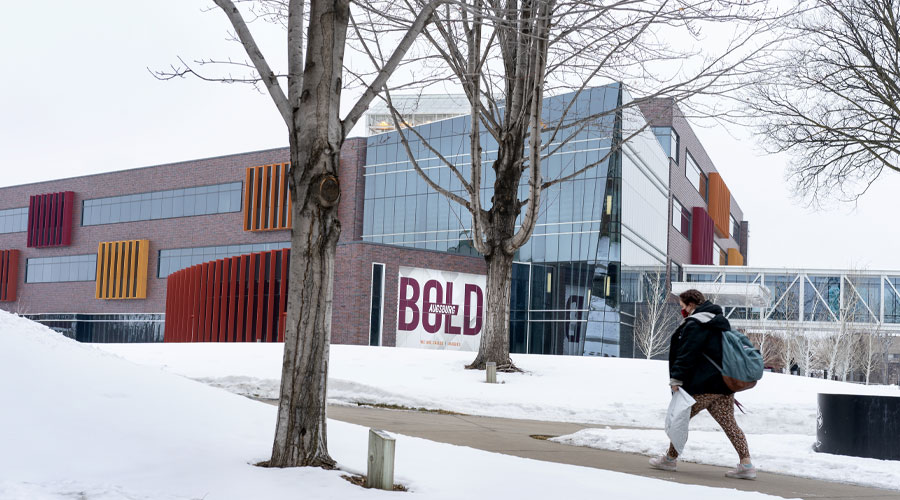 A student walking on the sidewalk in front of Hagfors Center with snow on the ground.