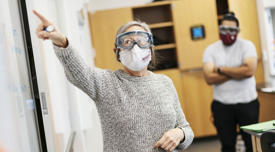 Professor teaching at the front of a science class with a face mask and eye wear on.