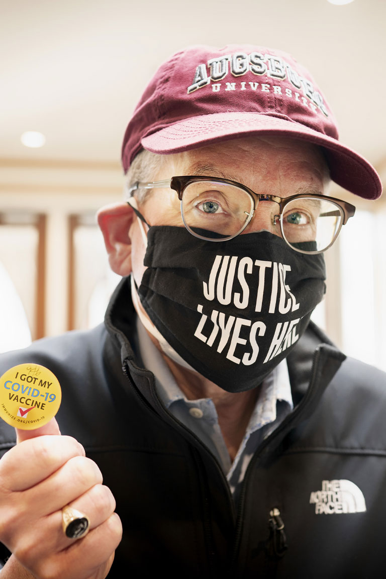 Augsburg President, Paul C. Pribbenow holding his "I Got My COVID-19 Vaccination" sticker. His face mask says "Justic Lives Here".