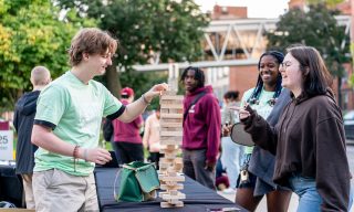 People playing a game of giant Jenga outdoors, with others watching and smiling. 