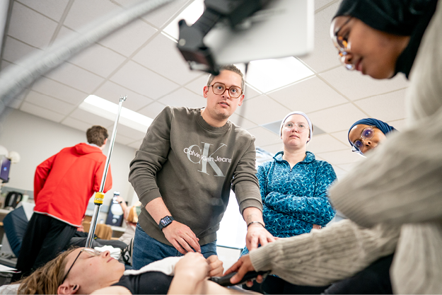 Group of people in a room focused on an individual lying down, with medical equipment nearby.