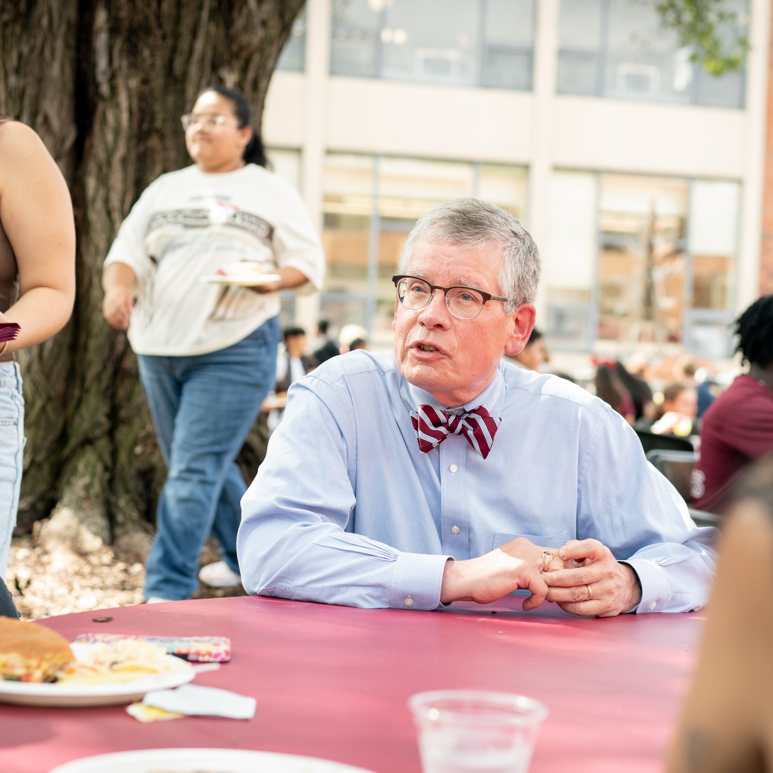 Paul Pribbenow, wearing a blue shirt and a striped bow tie, speaks with students at a table in the Quad during the opening convocation luncheon in 2024.