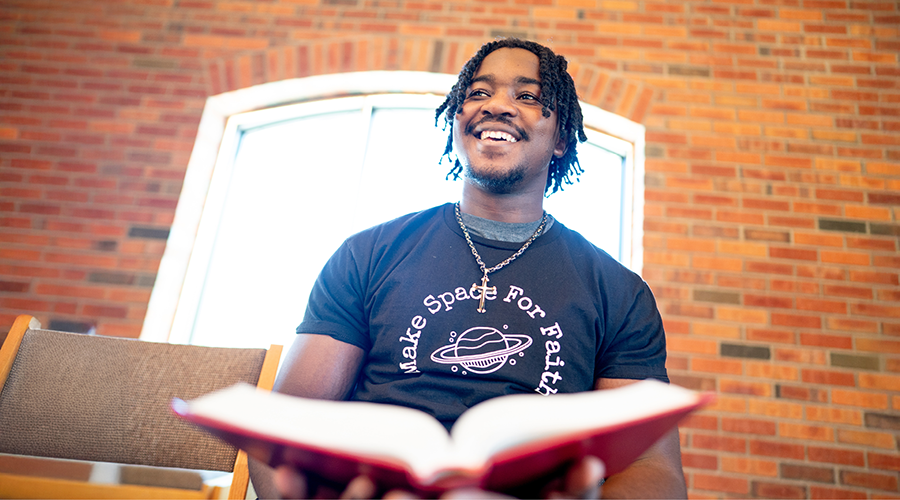Person smiling, wearing a black t-shirt with "Make Space For Faith" design, holding an open red book in a room with a brick wall and large window.