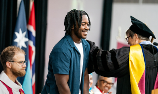 A young man on stage with people in academic regalia. Flags are visible in the background.