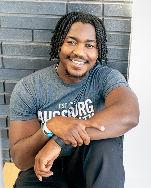 A man with dreadlocks sits on a bench, looking thoughtfully into the distance.