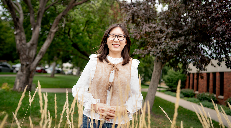 A woman wearing glasses stands in front of tall grass, enjoying the outdoors on a sunny day.