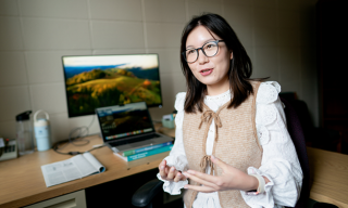 A woman with glasses is seated at a desk, with a computer behind her.