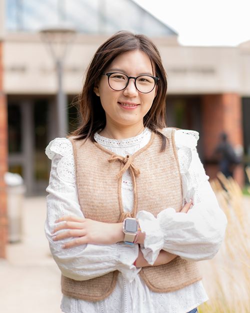 A woman wearing glasses and a vest stands in front of a building, smiling confidently.