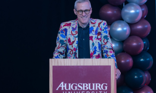 Person speaking at a podium with a colorful jacket and balloons in the background.