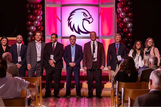 A group of nine people stand on stage in formal attire with an eagle silhouette in the background and balloon columns on the sides.