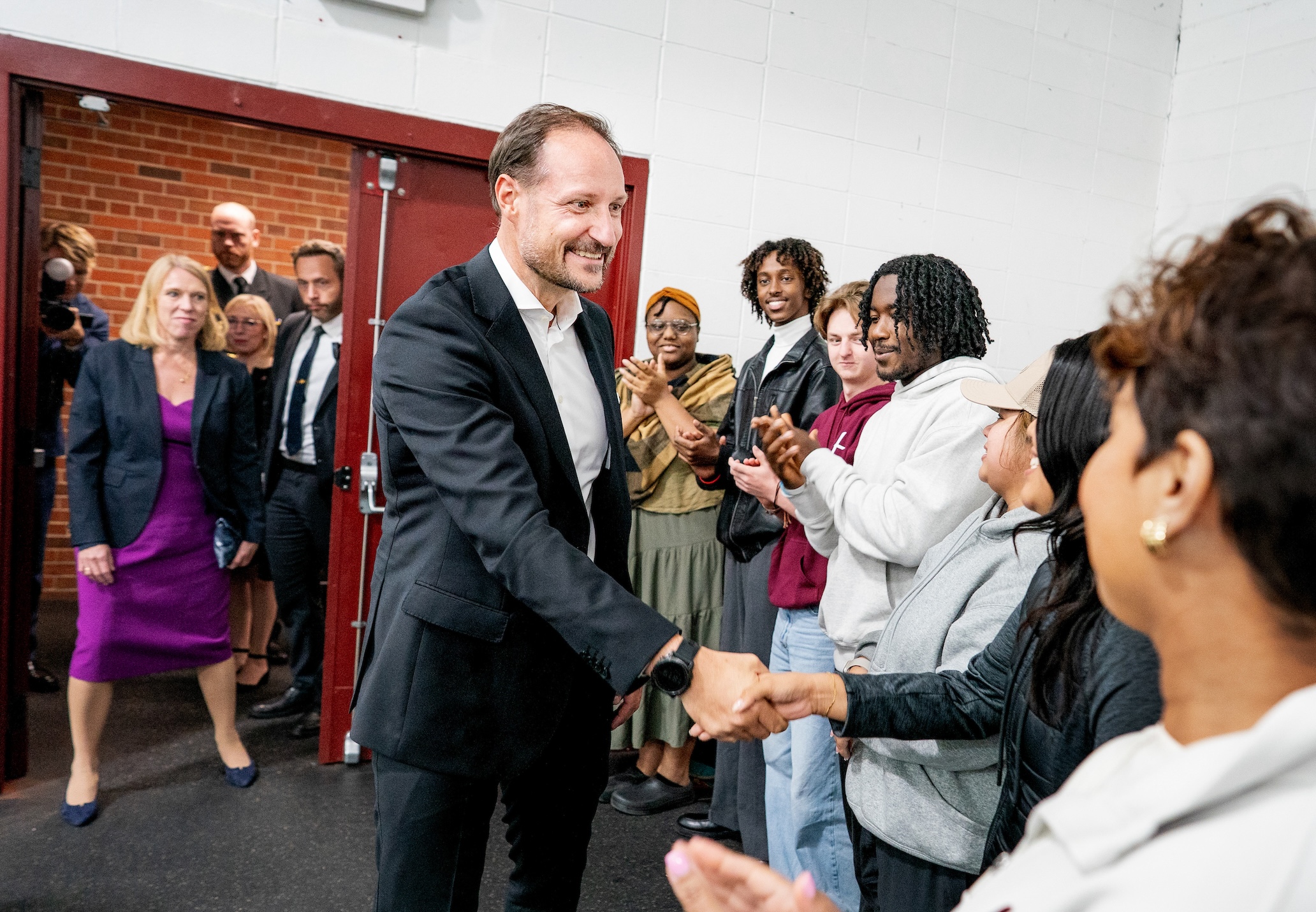 His Royal Highness Crown Prince of Norway, wearing a dark suit and white shirt, greets a smiling line of Augsburg students and staff in the ice rink.