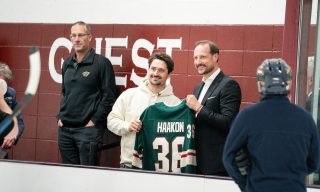 His Royal Highness Crown Prince Haakon of Norway and Minnesota Wild hockey player Mats Zuccarello stand next to each other, smiling and each holding one sleeve of a Wild jersey with "Haakon" on the back. 