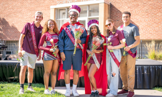 A group of six people in front of Science Hall, wearing maroon sashes and holding flowers.