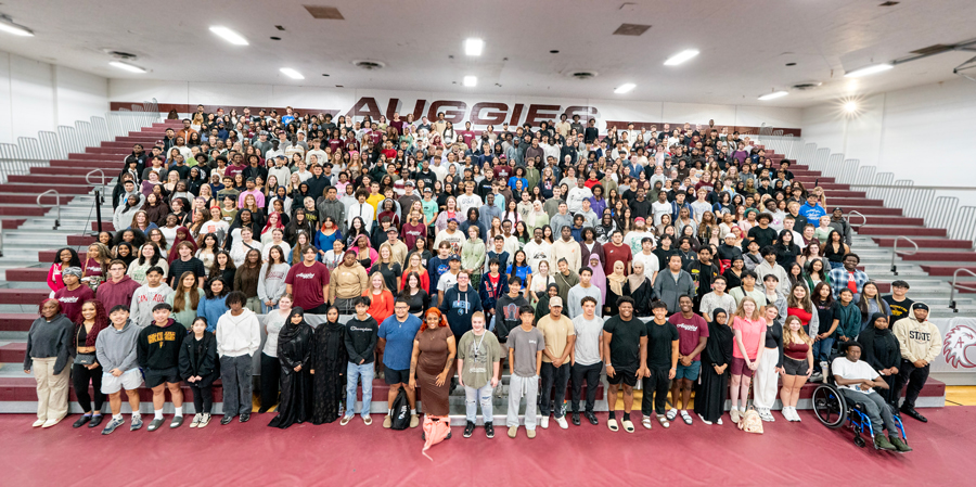 A large, diverse group of people stands on bleachers in a gym with "AUGGIE" in the background.