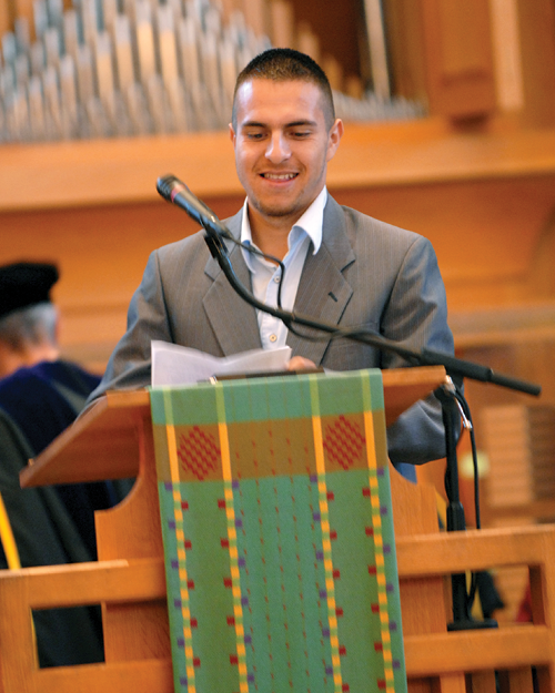 A man in a grey suit speaks at a podium decorated with a colorful woven cloth.