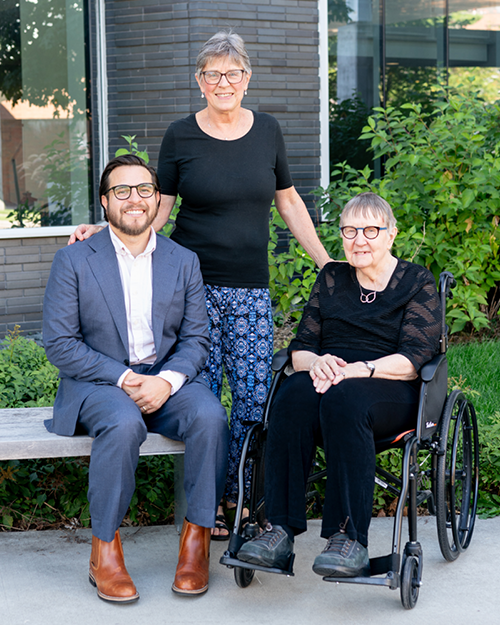 Three people, two seated and one standing, outdoors by a modern building.