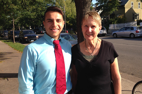 "Man in a blue shirt and red tie standing next to a woman in a black top on a sidewalk with trees and parked cars in the background."