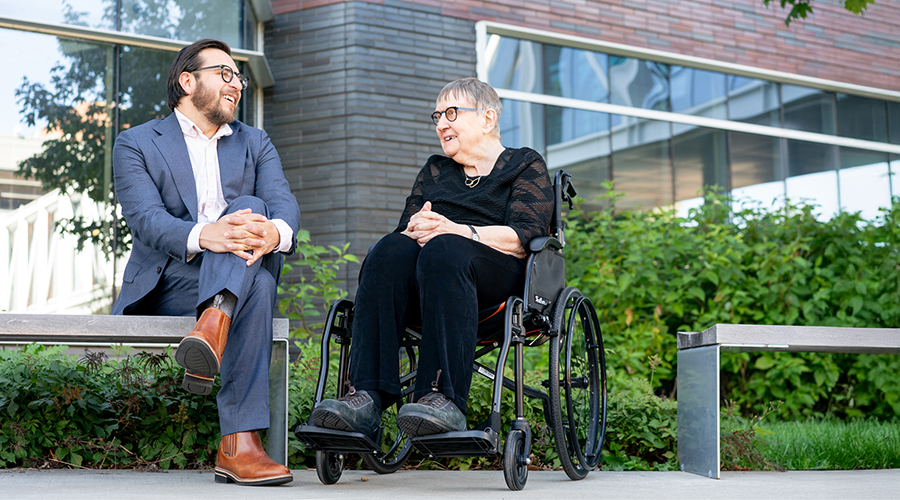 A man in a blue suit and a woman in a wheelchair converse outdoors on a sunny day.