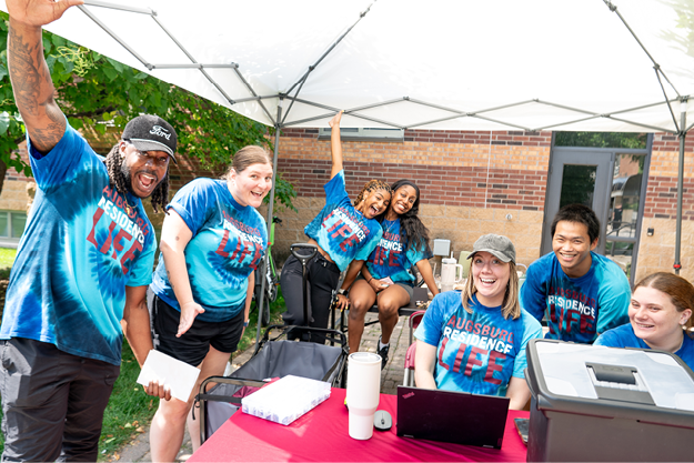 A group of smiling people wearing blue tie-dye shirts gather under a canopy with a laptop on a table.