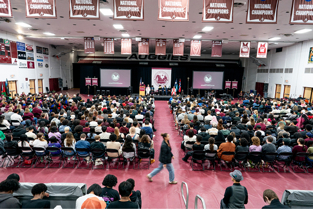 A packed auditorium with an audience facing a stage with a speaker, banners, and flags.