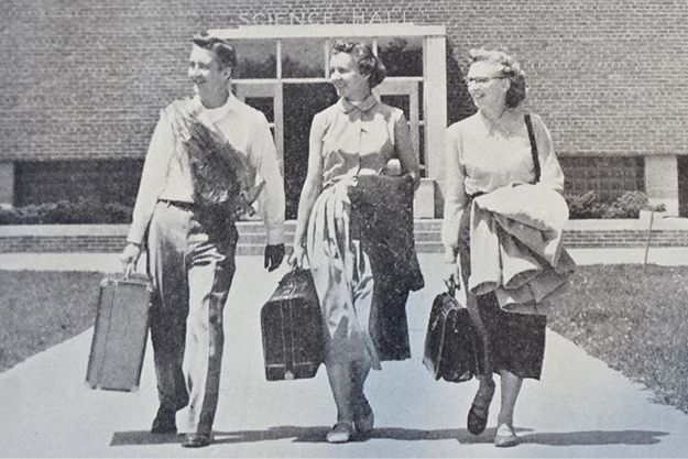 Three people walking with suitcases in front of a building labeled "Science Hall."