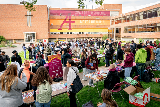 A crowded university event with students gathered around tables in front of a building with a large sign.
